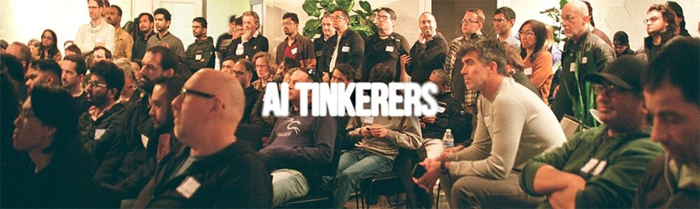 (Photo) This photo shows a large group of people seated and standing, likely attending a conference or meeting. The text 'AI TINKERERS' is superimposed over the image. Text: AI TINKERERS group of people | indoor | large crowd, superimposed text | candid | Colors: #4A4A4A, #A9A9A9, #FFFFFF Note: The image depicts a real-world scene with people, which is characteristic of a photograph. The text is added on top of the photo.