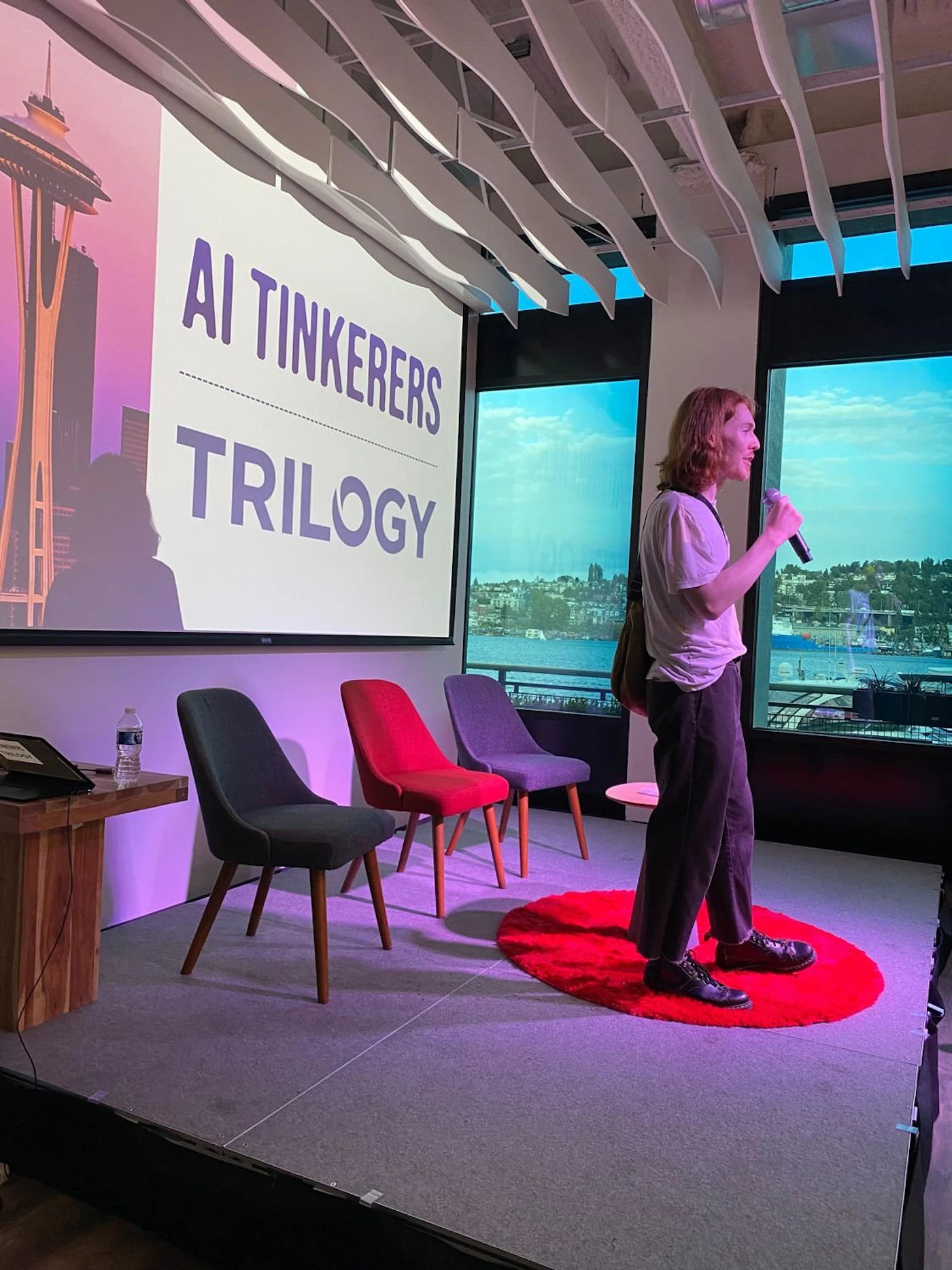 (Photo) A person with a microphone stands on a small red rug on a stage, in front of a projected banner and some chairs. There is a view of a cityscape through windows behind them. Text: AI Tinkerers TRILOGY person | indoor | presentation, stage, audience chairs, projected banner | candid Note: This is a real-life captured image of a person presenting, making it a photograph. It contains a projected banner, but the primary subject is the scene itself.