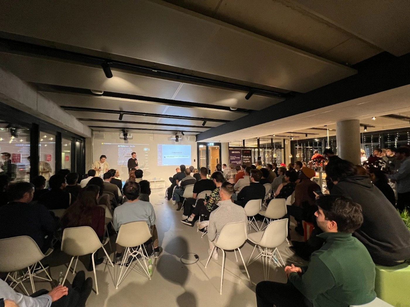 (Photo) A photo of an indoor presentation or lecture, with an audience seated in chairs facing a screen and two speakers. people | indoor | presentation, audience, speakers | candid Note: The image is a real-life captured image of a scene with people and a presentation, making it a photograph.