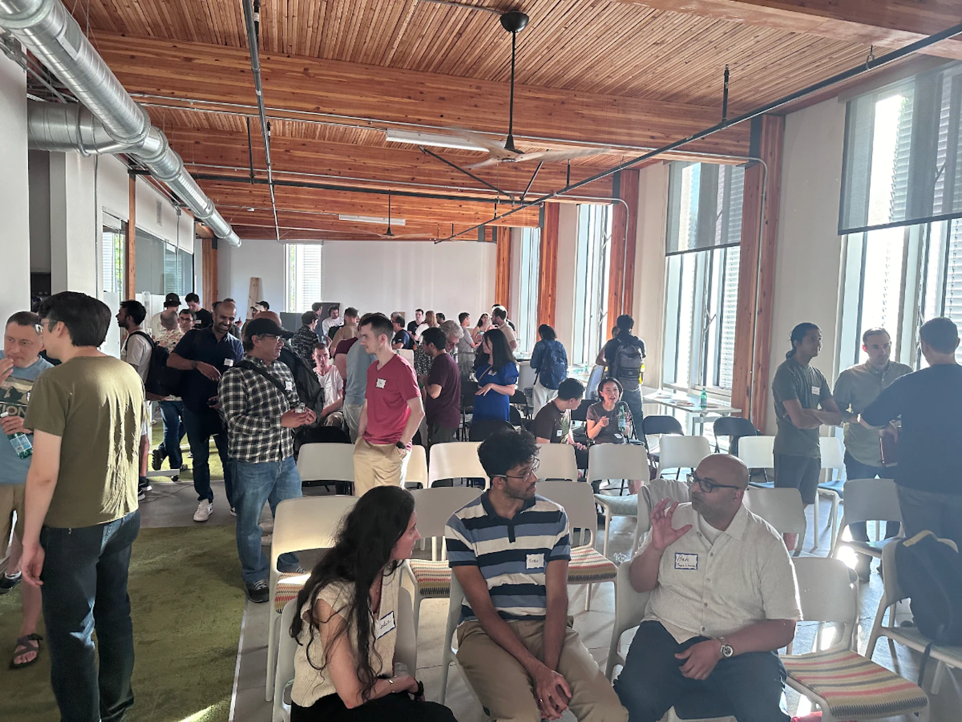 (Photo) A photo of a group of people gathered in a large room, possibly at a conference or networking event. people | indoor | large group, casual setting, some people seated, some standing | candid Note: The image is a real-life capture of a scene with people, indicating it's a photograph rather than an illustration or other image type.  It's not a logo or banner, and there's no discernible handwriting.