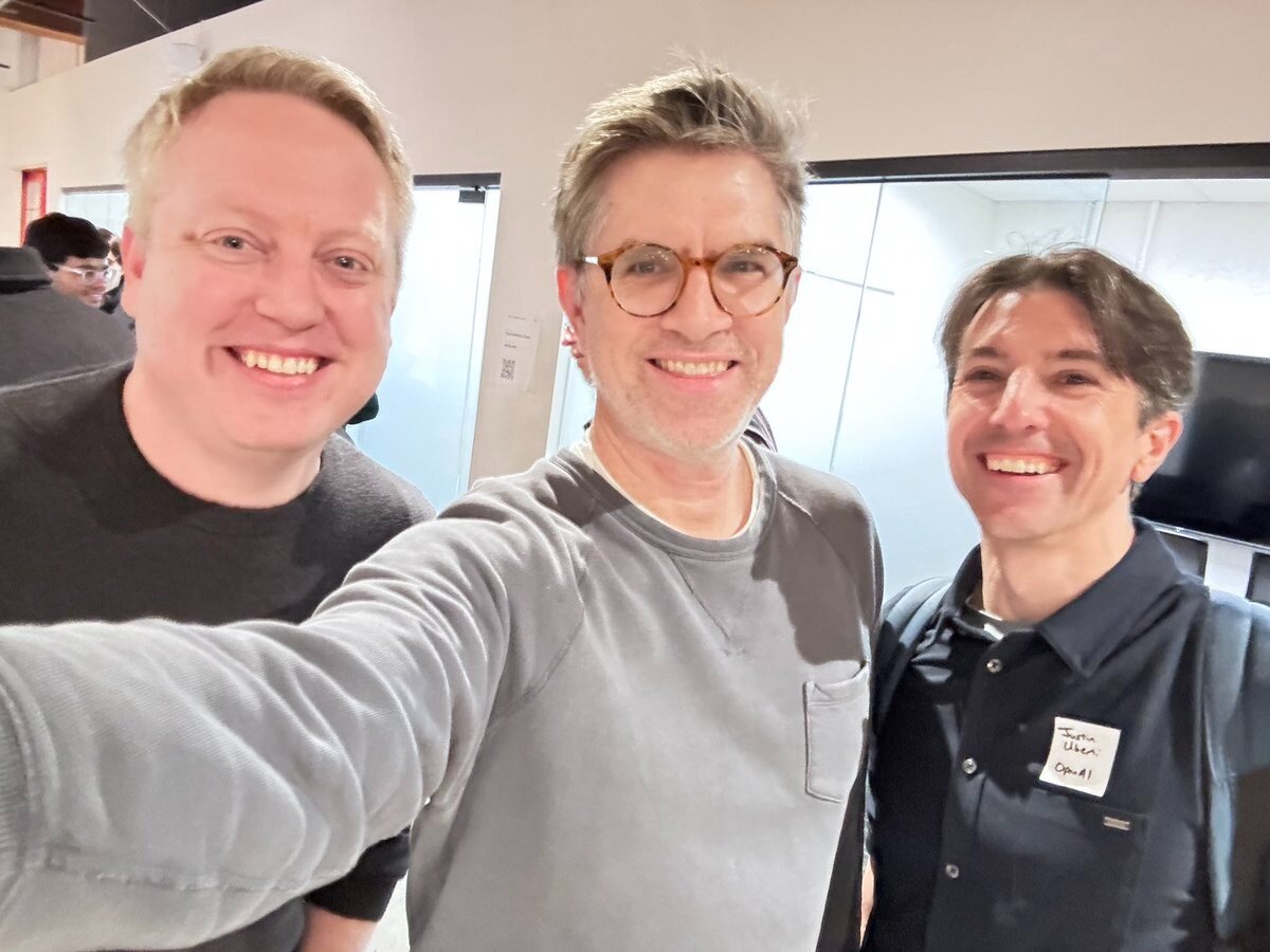 (Photo) Three men are posing for a photo indoors. They are all smiling at the camera. Text: Justin Uber OpEd! people | indoor | multiple people, smiling | #D3D3D3 | #000000 | #FFFFFF | candid | Colors: #D3D3D3, #000000, #FFFFFF Note: The image shows a real-life scene with people, and the style is consistent with a photograph. There is also some handwritten text on a badge.