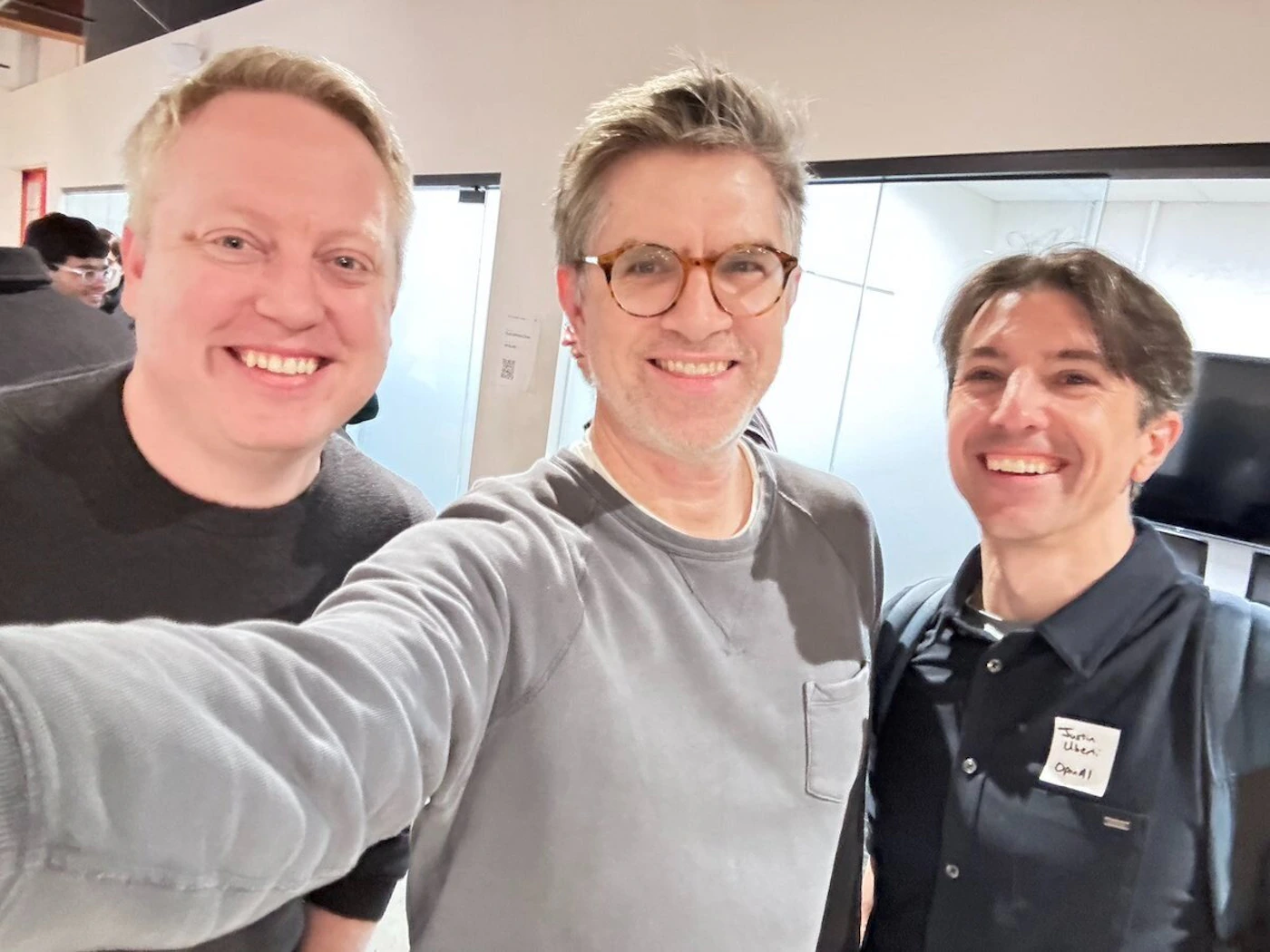 (Photo) Three men are posing for a photo indoors. They are all smiling at the camera. Text: Justin Uber OpEd! people | indoor | multiple people, smiling | candid | Colors: #D3D3D3, #000000, #FFFFFF Note: The image shows a real-life scene with people, and the style is consistent with a photograph. There is also some handwritten text on a badge.