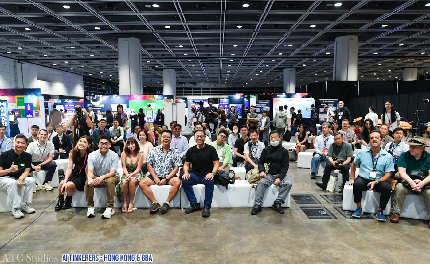 (Photo) A wide-angle photograph captures a large group of people, primarily adults, posing for a group shot at an indoor event or conference. They are seated and standing in rows, facing the camera, with various exhibition booths visible in the brightly lit background. Text: Ali G Studios AI TINKERERS - HONG KONG & GBA A large group of people at an event | Indoor event hall or convention center | Many individuals of diverse ages and appearances, event booths/displays in the background, high grid-patterned ceiling, large open space. | Group portrait, wide shot | Colors: #8C8C8C, #E0E0E0, #000000, #4A90E2, #9B59B6 Note: The image is a direct capture of real-world subjects (a large group of people and an event space) using photographic equipment, making 'photo' the most appropriate category.