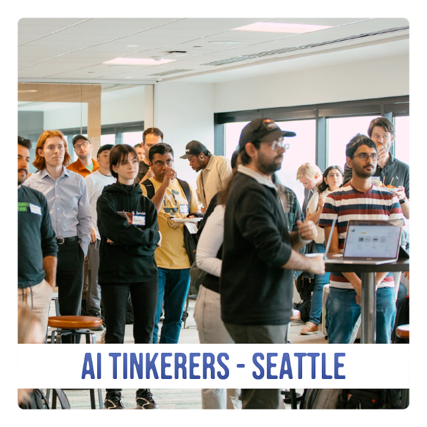 (Photo) A photograph of a group of people gathered in a large, modern room, seemingly at a conference or workshop. Text: AI TINKERERS - SEATTLE people | indoor | group of people, conference/workshop setting | candid Note: The image is a real-life captured scene of people in an indoor setting, making it a photograph. The text banner at the bottom provides context.