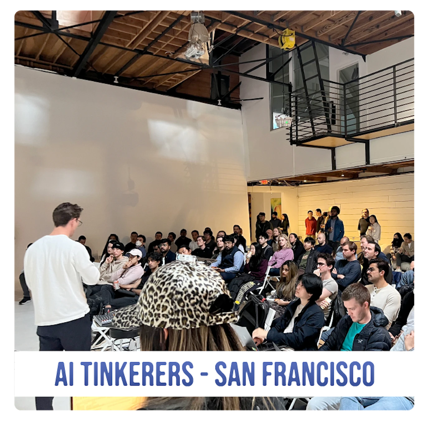 (Photo) A photo of a presentation or talk being given to a seated audience in a large, industrial-style room. Text: AI TINKERERS - SAN FRANCISCO people | indoor | presentation, audience, industrial venue | candid Note: The image is a real-life captured image of an event, showing a speaker and an audience in a specific venue.  This makes it a photograph.