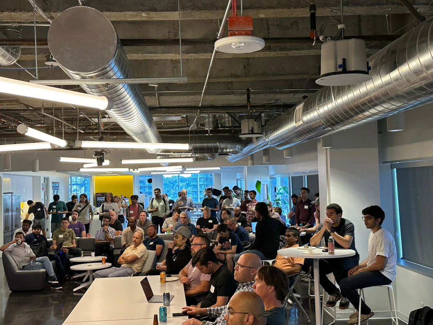 (Photo) A photo of a large group of people gathered in a modern office space, some seated and some standing, possibly for a presentation or casual meeting. people | indoor | large group, office setting, presentation or meeting | candid Note: The image is a real-life captured scene of people in an office environment, with no text or artistic elements suggesting other categories.