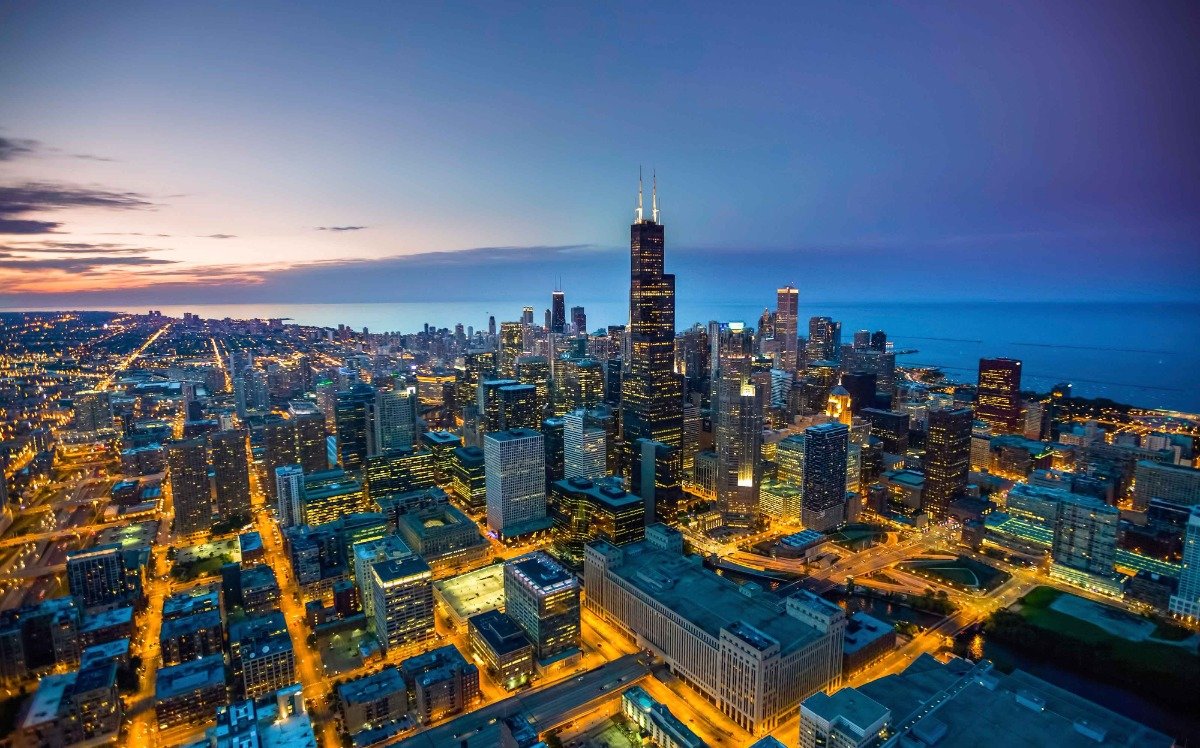 An aerial view of a cityscape at dusk, with lights illuminating the buildings and streets. The city is situated next to a body of water, possibly a lake or ocean.