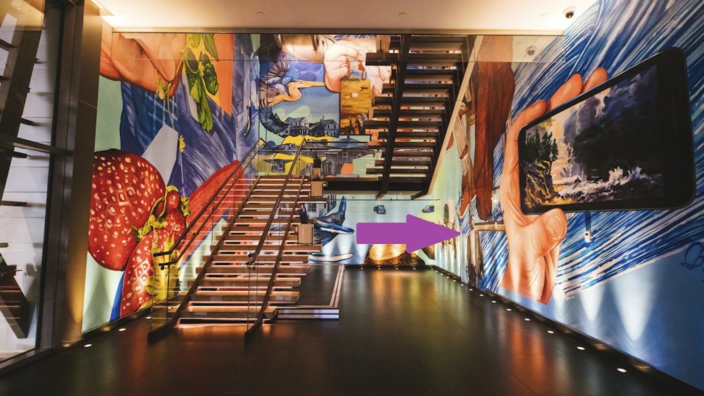 (Photo) A photo of an interior space with a staircase and walls adorned with a large, colorful mural featuring various images, including strawberries, a hand holding a phone displaying a landscape, and a whale. Interior space with mural | Indoor | Staircase, mural with various images (strawberries, hand holding phone, whale) | Architectural photography | Colors: #FF0000, #0000FF, #A0522D Note: The image is a photograph of an indoor scene, capturing the architectural elements and a mural. It's not a logo, illustration, banner, or handwriting.