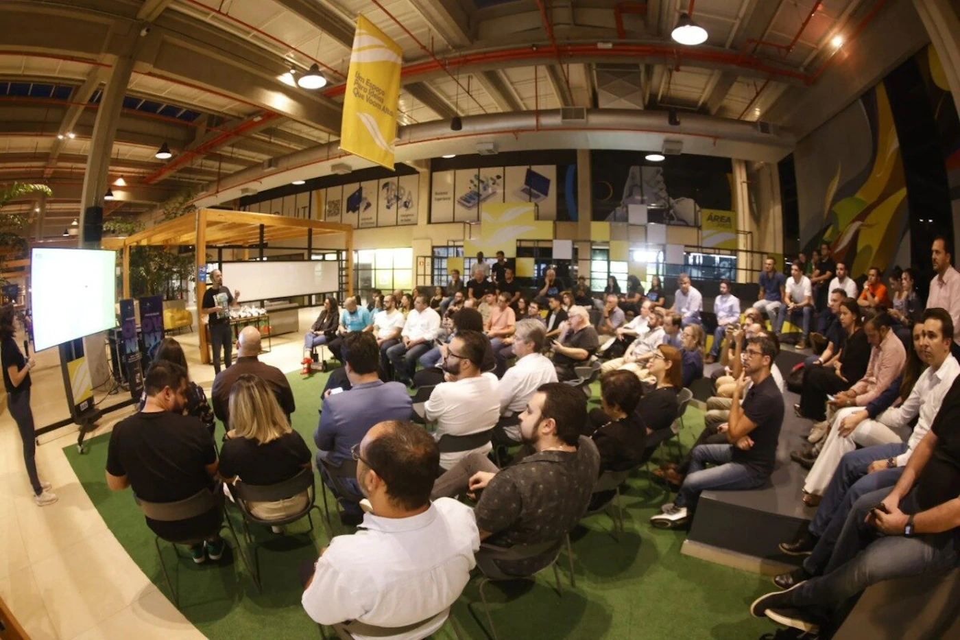 (Photo) A wide-angle photograph captures a large audience seated indoors, attending a presentation or event in a modern, industrial-style venue with high ceilings and exposed structure. Text: Um Espaço Pra Quem Voa Alto AREA Group of people attending a presentation/event | Indoor, large event space or co-working area | Large audience, two speakers, presentation screen, high industrial ceiling, yellow banner | Wide-angle/candid, event documentation | Colors: #FFFFFF, #000000, #FFD700, #3CB371 Note: The image is a documentary photograph capturing a real-world event with a large group of people in an indoor setting.