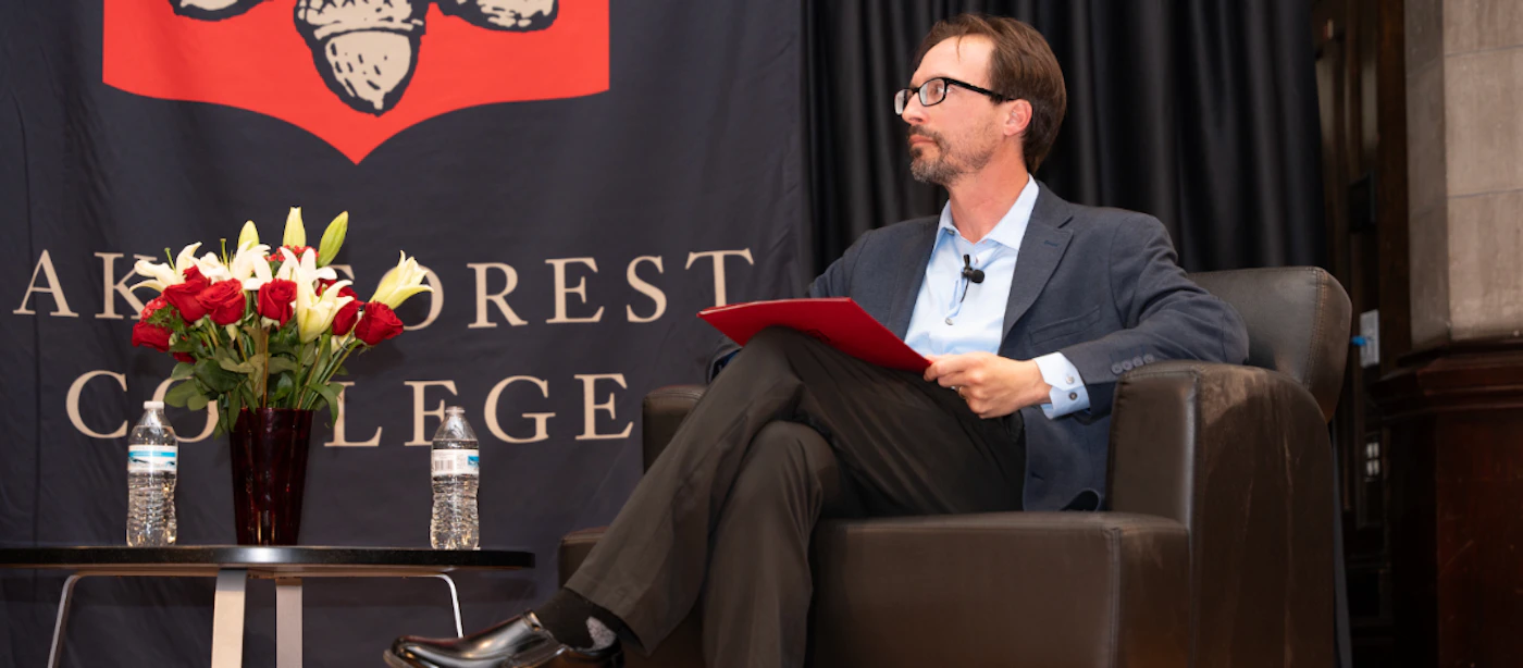 (Photo) A man in a dark suit and glasses sits in a brown leather armchair, holding a red folder and looking to the right. Behind him is a dark banner with a red and white logo and the text 'WAKE FOREST COLLEGE', with a vase of red roses and white lilies and two water bottles on a small table beside him. Text: WAKE FOREST COLLEGE person, event setting | indoor | man in a suit, banner with university logo and text, floral arrangement, water bottles, stage-like setup | candid | Colors: #26262b, #5c4438, #b80000, #e0e0e0 Note: The image is a realistic capture of a scene featuring a person, a banner, and various objects in an indoor event setting, characteristic of a photograph.