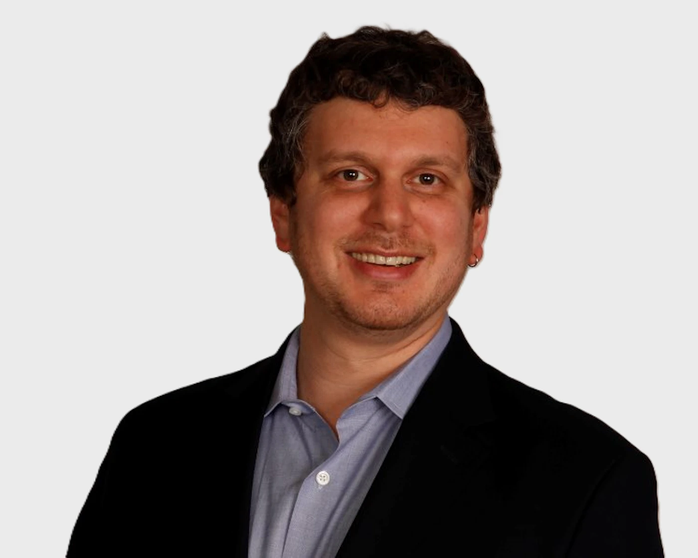 (Photo) A professional headshot of a smiling man with curly dark hair, wearing a black suit jacket and a light blue collared shirt, set against a plain light gray background. person (adult male) | indoor (studio/plain background) | professional attire, smiling expression, close-up portrait, small earring visible | portrait, headshot, professional | Colors: #000000, #E8E8E8, #336699, #A08060 Note: The image is a captured representation of a real-world subject (a person) using photographic techniques, typical of a professional portrait or headshot.