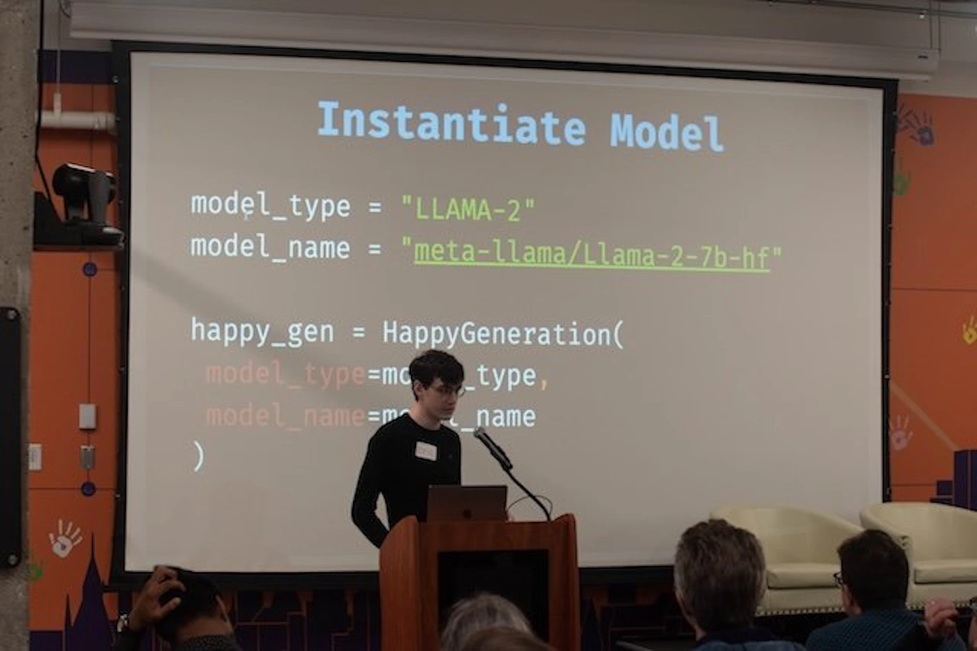 (Photo) A photo of a person presenting a slide about instantiating a large language model. Text: Instantiate Model model_type = "LLAMA-2" model_name = "meta-llama/Llama-2-7b-hf" happy_gen = HappyGeneration( model_type=mc _type, model_name=m^ name ) person | indoor | presentation, projector screen, audience | candid Note: The image is a photograph of a presentation, capturing a person, a projector screen, and an audience.  It's a real-world scene captured by a camera.