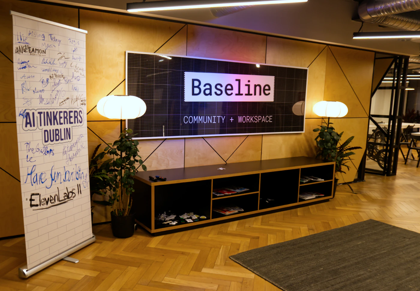 (Photo) A photograph of a modern workspace interior featuring a wooden wall with geometric paneling, above a low black shelving unit and a parquet floor. A large backlit sign displays the text "Baseline COMMUNITY + WORKSPACE," and a pull-up banner covered in handwritten signatures and messages stands to the left. Text: Baseline COMMUNITY + WORKSPACE AI TINKERERS DUBLIN The Builders Have fun building ElevenLabs!! Hip Aisling Telage Red S EAMON Woodin Happy C Aura The [unreadable] team [Numerous handwritten names/signatures/messages, largely unreadable] Interior architecture and event signage | indoor | Large backlit sign, geometric wooden wall paneling, signature banner, herringbone parquet floor. | candid | Colors: #A07F55, #1A1A1A, #FFFFFF Note: This is a photographic capture of an indoor architectural scene, documenting the interior design and signage of a modern co-working or event space.