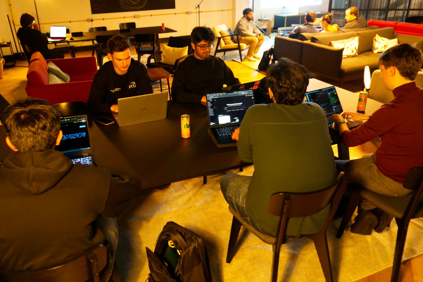 (Photo) A candid photograph captures a group of young adults intensely working on laptops around a dark table in a dimly lit, casual indoor lounge or co-working space, while other participants relax on couches in the background. Text: FCU clu [unreadable text on shirt] [unreadable text on screens] Group of people coding/collaborating | Indoor, casual lounge/co-working space | Multiple open laptops, warm yellow lighting, dark furniture | Candid, low-light | Colors: #333333, #8B4513, #FFD700 Note: The image is a photograph capturing a real-life scene of people engaged in an activity within an indoor setting, characteristic of a candid or documentary photo.