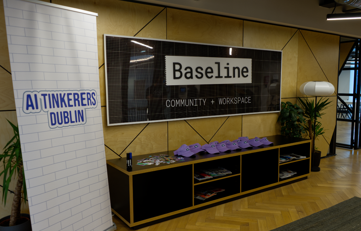 (Photo) A photograph of a modern indoor space, likely an office or event area, featuring a large wall-mounted sign for 'Baseline: COMMUNITY + WORKSPACE' and a pull-up banner for 'AI TINKERERS DUBLIN'. A long black cabinet with purple hats and various items is situated below the wall sign. Text: Baseline COMMUNITY + WORKSPACE AI TINKERERS DUBLIN interior office/event space, signage, furniture, plants | indoor, modern workspace | large wall sign, pull-up banner, long black cabinet, purple baseball caps, wooden wall paneling, herringbone floor, potted plants | candid, wide shot, well-lit | Colors: #B8860B, #000000, #FFFFFF, #8A2BE2, #228B22 Note: The provided image is a realistic capture of an indoor environment, typical of a photograph. It depicts a physical space with objects, furniture, and signage, rather than a graphic design, drawing, or handwritten document.