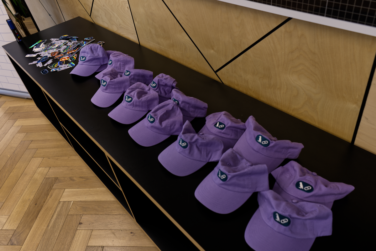 (Photo) A long black table displays a neat row of light purple baseball caps, each featuring a small, dark teal logo. To the left, a pile of colorful stickers is scattered on the table. The background shows a modern interior with light wood paneling featuring black geometric lines and a wooden herringbone floor. objects (baseball caps, stickers) | indoor (event space, office, or retail setting) | multiple identical caps with a distinct logo, a pile of assorted stickers, modern table and wall design, herringbone floor pattern | candid, product display | Colors: #A080D0, #000000, #D4B88C, #206060 Note: This image is a realistic depiction of physical objects (caps, stickers, furniture) in a real-world setting, characteristic of a photograph.