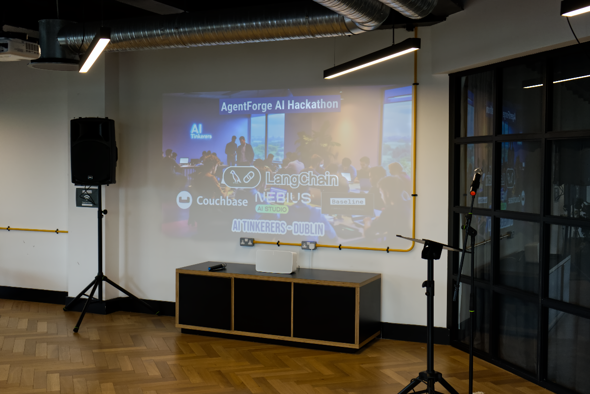 (Photo) An indoor photograph of a modern event space, featuring a large projection on a white wall displaying information about an 'AgentForge AI Hackathon'. The room is equipped with a speaker on a stand and a microphone on another stand, with a black cabinet positioned below the projection. Text: AgentForge AI Hackathon AI Tinkers Couchbase LangChain NEBIUS AI STUDIO Baseline AI TINKERERS-DUBLIN indoor event space, projected presentation, audio equipment | indoor | large projected screen, speaker, microphone, modern interior design with exposed ducts, wooden parquet floor | interior shot | Colors: #F5F5F5, #000000, #A07A4C, #FFD700, #4A206F Note: The image captures a real-world scene of an indoor event space, including equipment and a projected presentation, which fits the definition of a photograph.