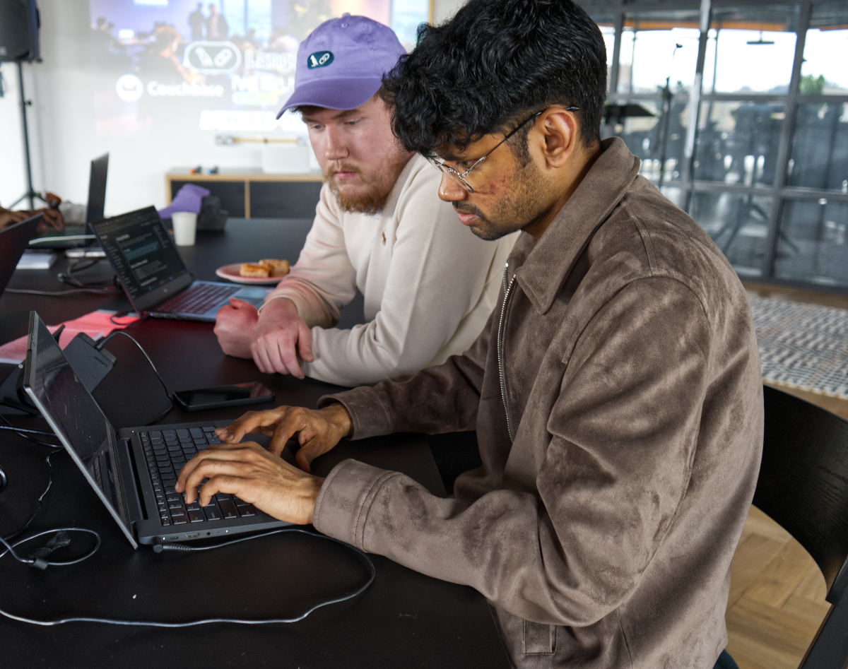 (Photo) The image captures two men in what appears to be a collaborative work setting. One man in the foreground is actively typing on a laptop, while another man in a purple cap is seated next to him, observing. Text: Couchbase Net Two men, technology, work/collaboration | Indoor, likely an office or co-working space with a projector screen and large windows in the background. | One man typing on a laptop, another observing, a projector displaying text and graphics, multiple laptops on a dark table, and some pastries. | Candid, observational, slightly informal. | Colors: #8C7360, #E0D7C9, #6A459E, #2C2C2C, #F0F0F0 Note: The image is a direct capture of a real-world scene featuring people and objects, characteristic of a photograph.
