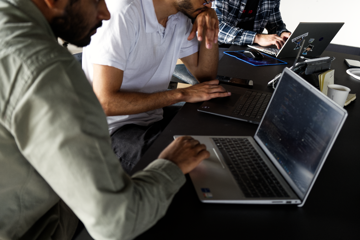 (Photo) A close-up photograph of three individuals, partially visible, sitting at a black table and working on their laptops. The scene appears to be an indoor office or co-working space, with a focus on the laptops and hands. Text: GitHub People working on laptops | Indoor, office or co-working space | Three partially visible individuals, multiple laptops, screens displaying code-like content, collaborative environment | Candid, documentary, slightly overhead | Colors: #000000, #FFFFFF, #8B947A, #2C3A4D, #A67C5E Note: The image is a realistic capture of a real-world scene featuring people and objects, characteristic of a photograph rather than a stylized illustration, logo, or banner.