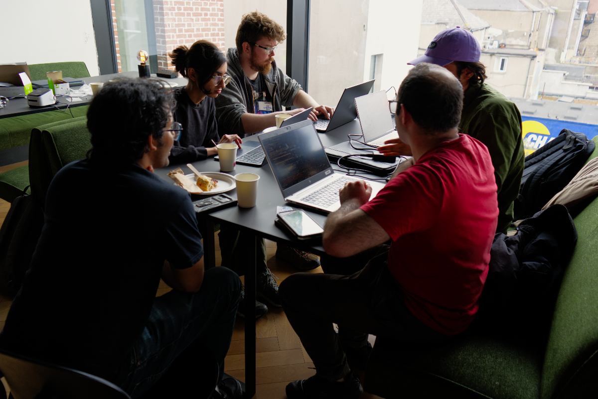 (Photo) An indoor photograph captures five individuals gathered around a black table, actively working on their laptops. The setting appears to be a collaborative workspace or cafe, with natural light streaming in from a large window that offers a view of an urban landscape. Text: CH[unreadable] Group of people working on laptops | Indoor workspace/cafe with an urban outdoor view | Five individuals, multiple laptops, food and drinks on table, large window, natural light | Candid, natural light, group shot | Colors: #2C2C2C, #386F38, #E73F3F, #C5A485, #A9B8BD Note: The image is a direct capture of a real-world scene featuring people and objects, characteristic of a photograph rather than an illustration, logo, or text-based image.