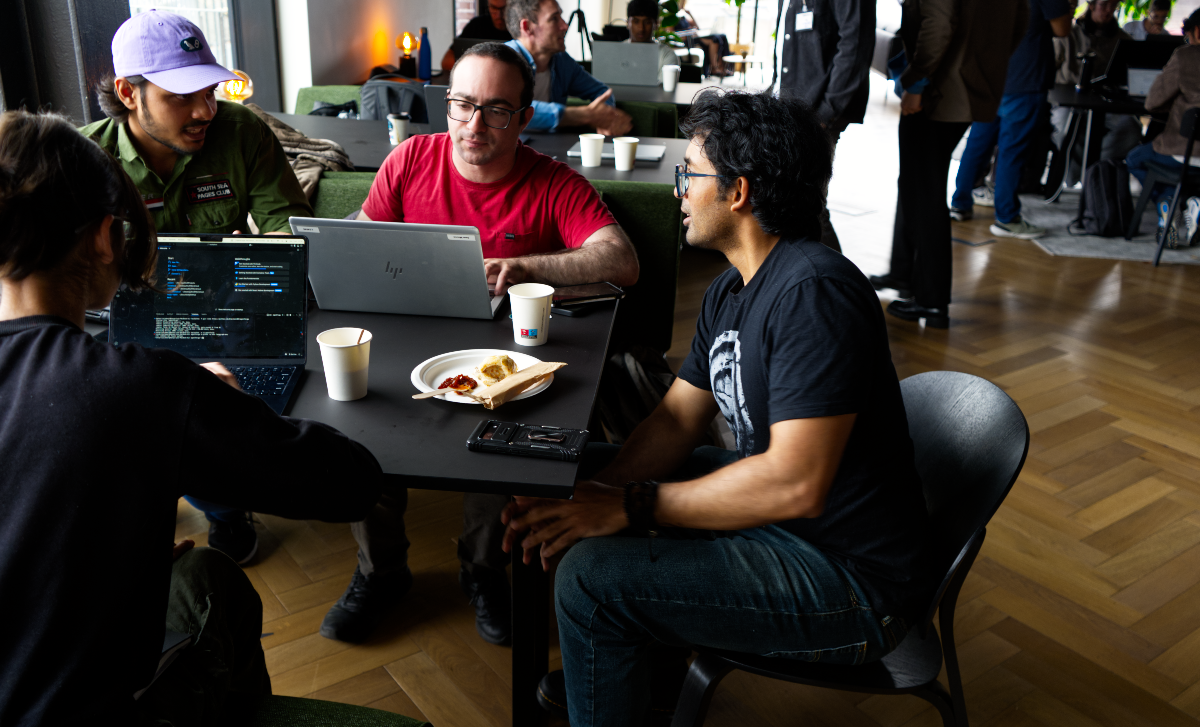 (Photo) A candid photograph captures a group of people, primarily men, engaged in discussion and working on laptops around a dark table in an indoor setting, possibly a co-working space or event. Text: SOUTH SUN PIZZA CLUB hp [unreadable] people, technology, food and drink | indoor, possibly a co-working space or casual meeting area | multiple people interacting, laptops, casual attire, wooden floor, bright ambient lighting | candid, realistic | Colors: #1A1A1A, #A6825C, #E03E3E, #8C6CE0 Note: The image is a realistic capture of a moment, showing people, objects, and an environment, which is characteristic of a photograph.