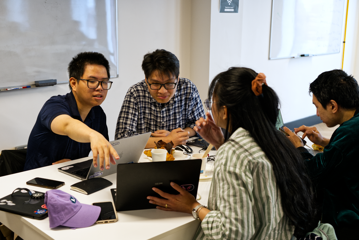 (Photo) A candid photo of four young adults gathered around a white table in an office or co-working space, engaged in a discussion or collaborative work with laptops and mobile devices. One person on the left is pointing towards a laptop screen, while others are focused on their devices or interacting. Text: Wireless [unreadable] People collaborating/working | Indoor, office/meeting room | Four individuals, multiple laptops and mobile phones, whiteboards in the background, group discussion/work, casual attire. | Candid, natural light, slightly warm tone | Colors: #FFFFFF, #1F3055, #4A6C8C, #8BA08A, #000000, #8D62C2, #B76D3E Note: The image is a direct capture of a real-world scene, featuring people and objects in a natural setting, which is characteristic of a photographic medium.