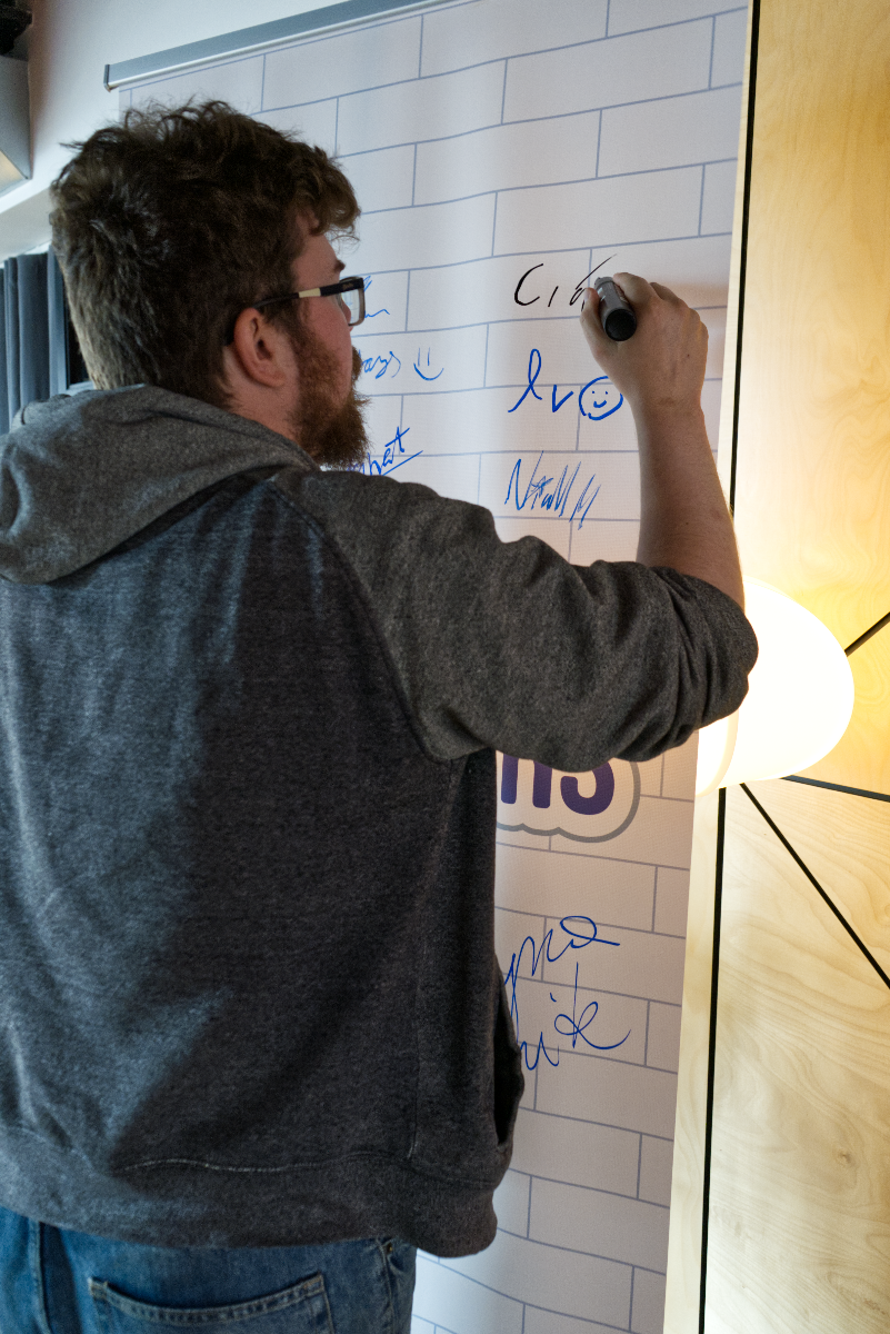 (Photo) A man with a beard and glasses is seen from behind, wearing a grey hoodie and blue jeans, writing on a large white board with a black marker. The board features a subtle brick-like pattern and is covered with various handwritten notes and drawings in blue and black ink. Text: Cia lv :) Niram [unreadable]ss :) [unreadable]hat mo mite IIS A person writing on a whiteboard/banner | Indoor, possibly an event space or office | Man with beard and glasses, large white board with brick pattern, various handwritten notes and drawings, modern light fixture, wooden wall paneling | Candid, observational | Colors: #666666, #336699, #FFFFFF, #000000, #A0522D, #FFD700 Note: This image is a direct photographic capture of a real-world scene, showing a person interacting with an object. It is not an illustration, logo, banner design, or a standalone piece of handwriting.