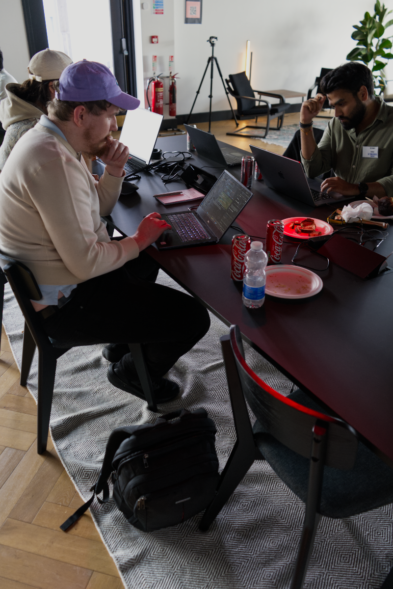 (Photo) A group of people are seated around a long, dark table in an indoor setting, actively engaged with their laptops. Drinks and snacks are scattered on the table, suggesting a collaborative work or study environment. Text: Coca-Cola people, objects (laptops, food, drinks) | indoor, office/co-working space | multiple people working on laptops, casual gathering, snacks and beverages on the table | candid, natural light | Colors: #1A1A1A, #F5F5F5, #D80000, #E0D4C5, #6B7B5B Note: The image is a realistic capture of a scene featuring people and objects in a real-world setting, which is characteristic of a photograph.