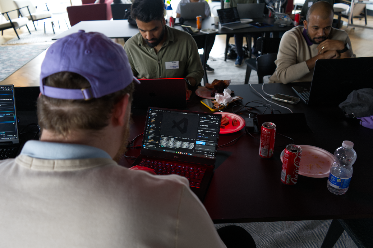 (Photo) The image captures an indoor scene of several individuals, primarily men, seated at dark tables, engrossed in working on their laptops. The foreground shows a person from behind wearing a purple cap, with a laptop displaying a code editor (VS Code), while other individuals are visible in the background also working on computers. Text: Coca-Cola ADAM [unreadable] Explorer Search Source Control Run and Debug Extensions Settings Account Manage People working on laptops | Indoor, likely an office or co-working space | Multiple people, laptops with coding interfaces, drinks (Coca-Cola cans, water bottle) and plates on tables, casual work environment, focus on the back of a person's head in the foreground. | Candid, eye-level, slightly overhead perspective | Colors: #201E1E, #D0C9B9, #8B6FE9, #E42222, #A9A9A9 Note: This image is a direct capture of a real-world scene featuring people and objects, characteristic of a photograph rather than an illustration, logo, or text-based banner.