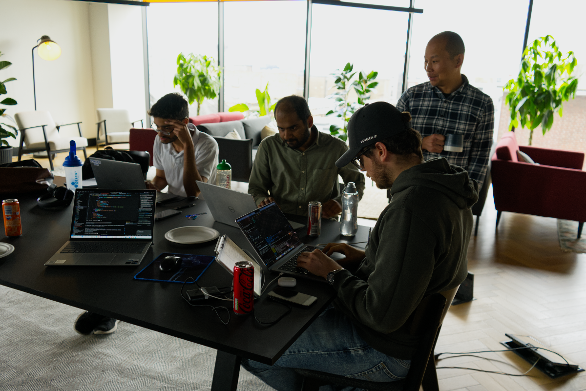 (Photo) A group of four men are gathered around a dark table in a bright, modern office or co-working space, actively working on their laptops. The room features large windows, various green plants, and lounge furniture in the background. Text: HYGOLF Coca-Cola Myprotein Gatorade [unreadable code] people working | indoor, modern office/co-working space | multiple people, laptops, large windows, plants, lounge area | candid, natural light | Colors: #1A1A1A, #4CAF50, #D2B48C, #F5F5F5, #B71C1C Note: The image is a realistic capture of people and an environment, consistent with a photograph. It depicts a scene rather than a graphic design, logo, or handwritten content.