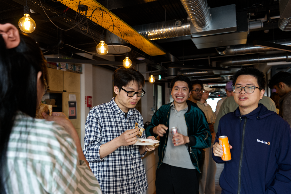 (Photo) A candid indoor photograph captures a group of people, primarily young men, socializing. The foreground features three men, one eating from a plate, another smiling and holding a can, and a third holding an orange can, all under industrial-style lighting with exposed ducts. Text: Reebok group of people | indoor, likely a casual office or event space | multiple people, social gathering, industrial-style decor, some people eating and drinking | candid, group shot | Colors: #2B3B5E, #FF9900, #FFD700, #2F6B58 Note: This image is a realistic depiction captured by a camera, showing people in an indoor environment, characteristic of a photograph.