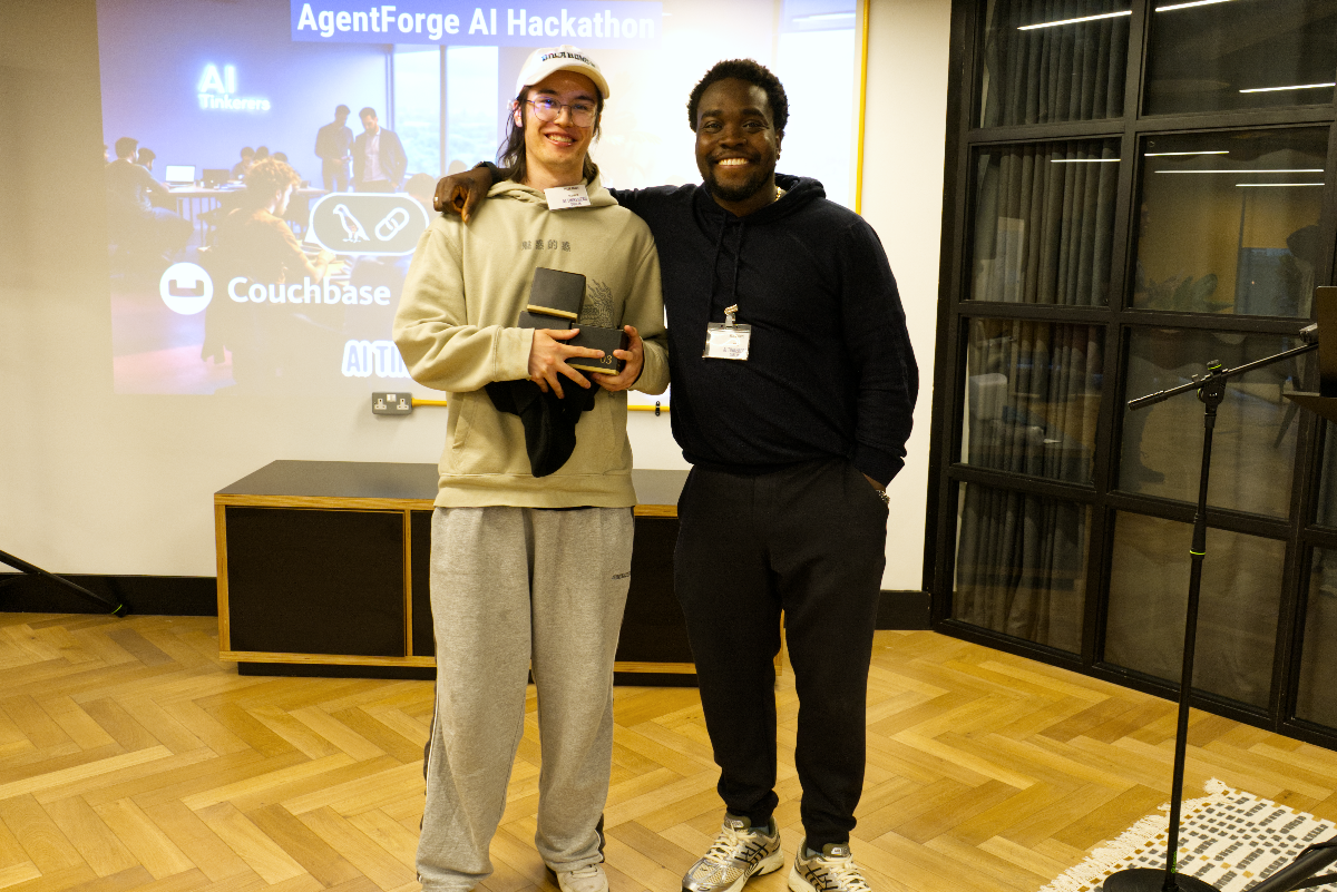 (Photo) The image shows two men smiling and standing side-by-side in an indoor setting, likely at an event. The man on the left is holding a black and gold award, while a projector screen in the background displays text related to an 'AgentForge AI Hackathon'. Text: AgentForge AI Hackathon AI Tinkers Couchbase AI TI[unreadable] 知行合一 AgentForge Two men, one holding an award, at an event. | Indoor event space or conference room. | Projector screen displaying event information, wooden herringbone floor, large black-framed windows, and a microphone stand. | Event photography, candid. | Colors: #A87B4A, #2C2C2C, #E0E0E0, #808080 Note: This image is a photograph capturing a real-world scene with people and objects, rather than a drawing, logo, or text-heavy banner. It depicts an event or moment in time.