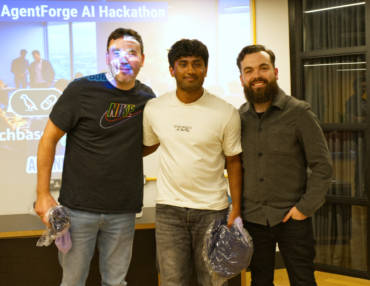 (Photo) A photograph capturing three men standing side-by-side, smiling at the camera, in an indoor setting. A projector screen behind them displays text related to an 'AgentForge AI Hackathon' and other partial information. Text: AgentForge AI Hackathon NIKE FUTURE PROJECTS chbas A N Three men | Indoor, likely an event space or office with a projector screen | Three men posing together; one wearing a Nike shirt, another a 'Future Projects' shirt; a projector screen with event details and icons in the background. | Candid/event photo | Colors: #2A2A2A, #F0F0F0, #A07F60, #8098B0 Note: This image is a direct capture of real-world subjects and their environment, characteristic of a photograph. It shows people posing and an event setting.