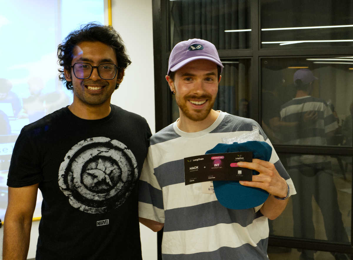 (Photo) Two smiling men are posing for a photo indoors. The man on the left wears a black t-shirt with a distressed Captain America shield logo, while the man on the right, wearing a striped shirt and a purple cap, holds a blue cap and a black event card. Text: MARVEL LangChain HACK INT HACKATHON PRIZE ALTITUDE DEV people | indoor | two men, smiling, one holding an event card and cap, Captain America shield logo on t-shirt, reflections in glass partition | candid, event photo | Colors: #1A1A1A, #F0F0F0, #A0B0C0, #9E8ED2, #007B8C Note: The image is a photographic capture of real people in a real-world setting, depicting a moment or event rather than a graphic design, illustration, or text document.