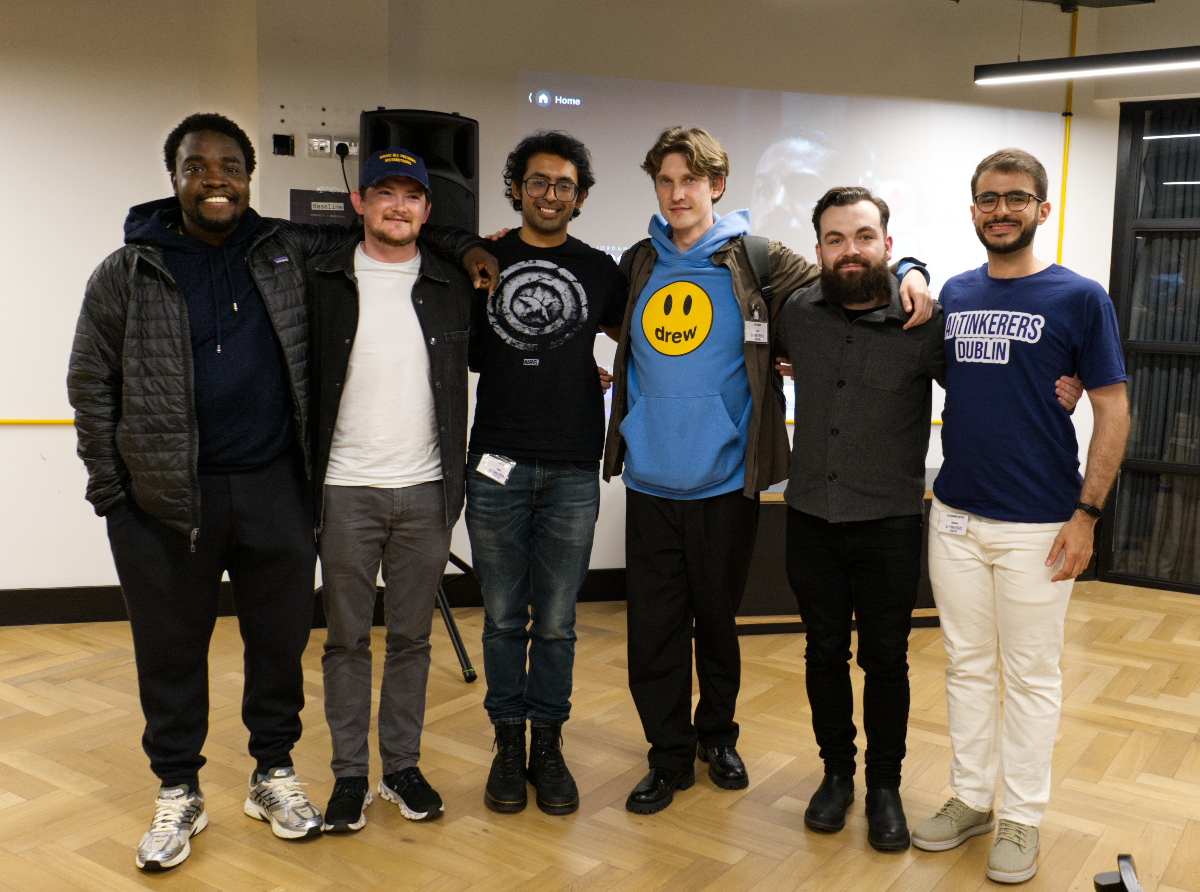 (Photo) A group of six men are standing together in an indoor setting, smiling and posing for a photo with their arms around each other's shoulders. A projector screen is visible in the background. Text: Home drew AI TINKERERS DUBLIN [unreadable] group of people | indoor | six men, smiling, arms around shoulders, projector screen in background, casual attire | group portrait, candid | Colors: #000000, #1A2B4D, #66CCFF, #F5F5F5, #D2B48C Note: The image captures a real-life scene with people and a physical environment, which is characteristic of a photograph. It is not an illustration, logo, banner, or handwriting.