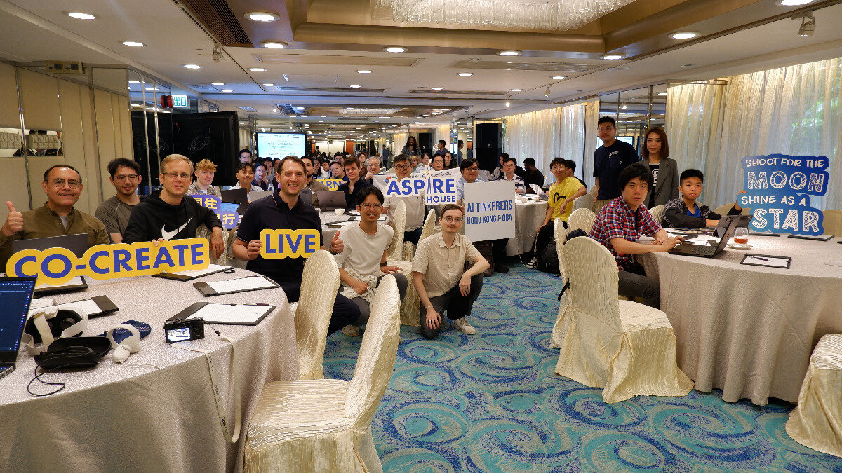 A large group of people gathered in a banquet hall for a tech-related event, with many individuals holding up motivational signs.. Text: CO-CREATE LIVE ASPIRE HOUSE AI TINKERERS HONG KONG & GBA SHOOT FOR THE MOON SHINE AS A STAR