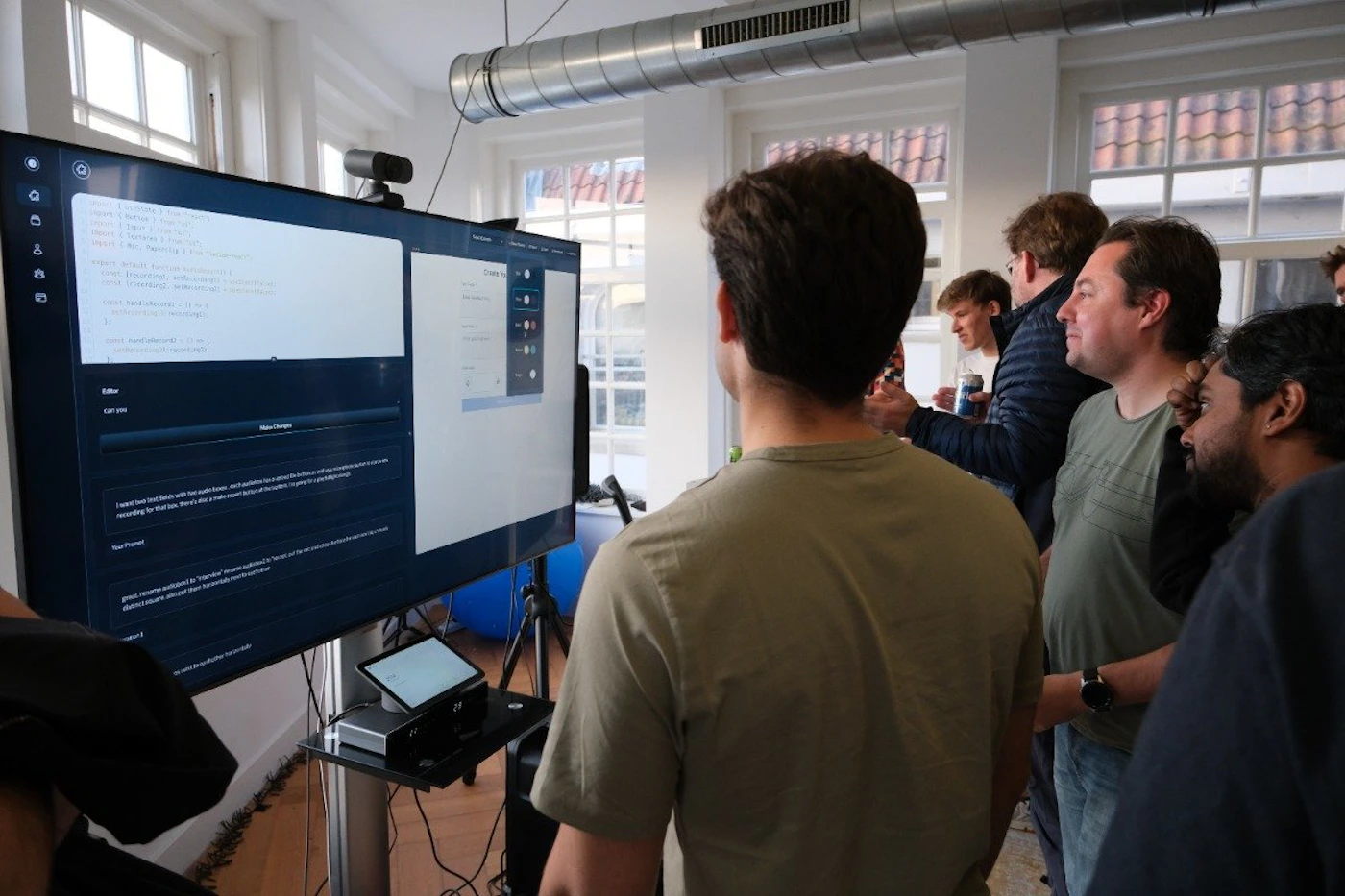 (Photo) A group of people are gathered in a room, looking at a large screen displaying code and a user interface. people | indoor | group of people, computer screen, code display | candid Note: The image is a real-life captured scene of people in a room, making it a photograph. It's not a staged image or artwork.