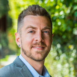 (Photo) A headshot of a man with short, styled hair and a beard, wearing a gray blazer and light blue shirt. He is smiling slightly and looking directly at the camera. person | outdoor | headshot, smiling | portrait Note: The image is a real-life photograph of a person, likely a professional headshot.