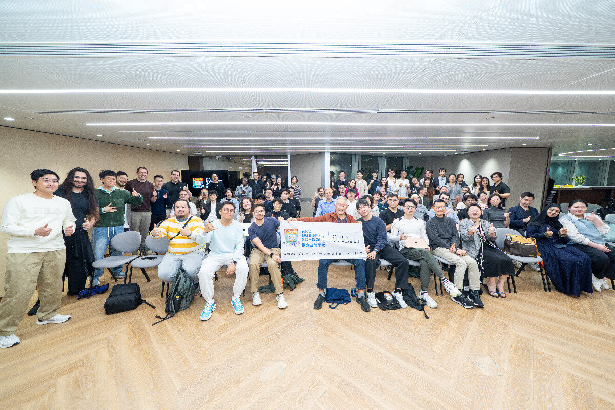 A large group of students and staff from HKU Business School posing for a group photo in a modern indoor hall, with some individuals holding a promotional banner.. Text: HKU BUSINESS SCHOOL 港大經管學院 MASTERS PROGRAMMES Career Development and Training (CDT) FILA