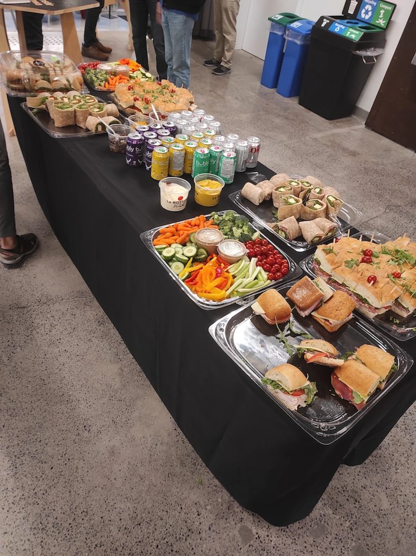 (Photo) A table laden with a variety of food items, likely for a catered event or gathering.  There are sandwiches, wraps, vegetables with dip, and canned drinks. Text: bubly food | indoor | catered food, buffet style, variety of options | candid Note: This is a real-life image captured with a camera, depicting a scene of food laid out for an event.  It's clearly a photograph of a buffet-style setup.