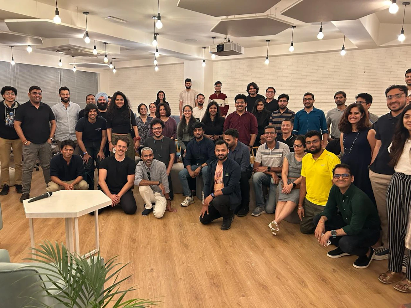 (Photo) A group of people are gathered together in a room, possibly for a meeting or a social event. They are posing for a photo, some standing and some sitting or kneeling. people | indoor | group photo | candid Note: The image is a real-life capture of a group of people in an indoor setting, indicating it's a photograph. There are no artistic elements suggesting illustration or other visual media.