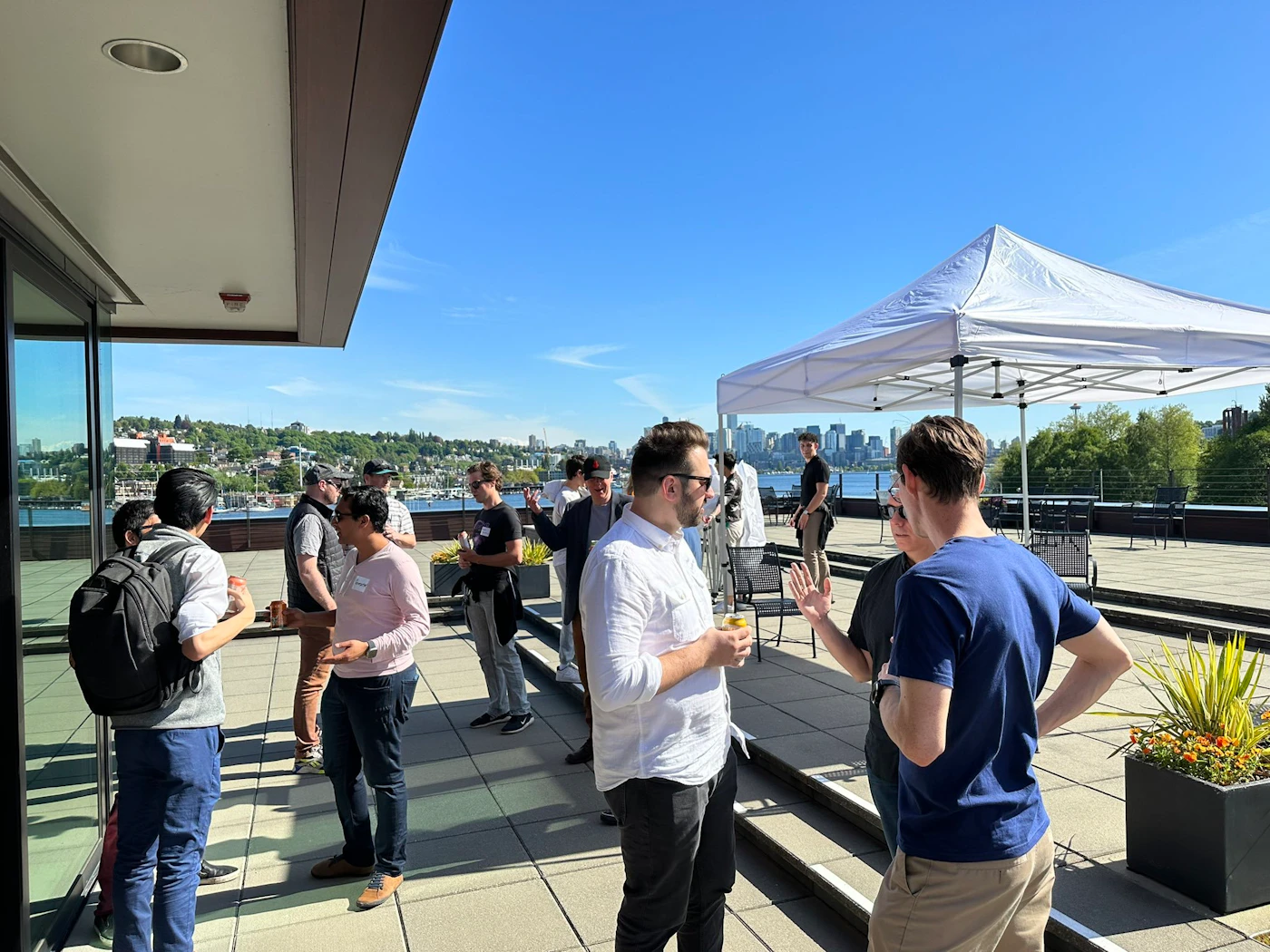 (Photo) A group of people are gathered on a rooftop patio, some are talking in small groups while others stand individually.  A white canopy tent is set up on the patio and a body of water and cityscape are visible in the background. people | outdoor | cityscape, rooftop patio, group of people | candid Note: This image is a real-life captured image of people in a setting, making it a photograph. It is not a drawn or computer-generated image.