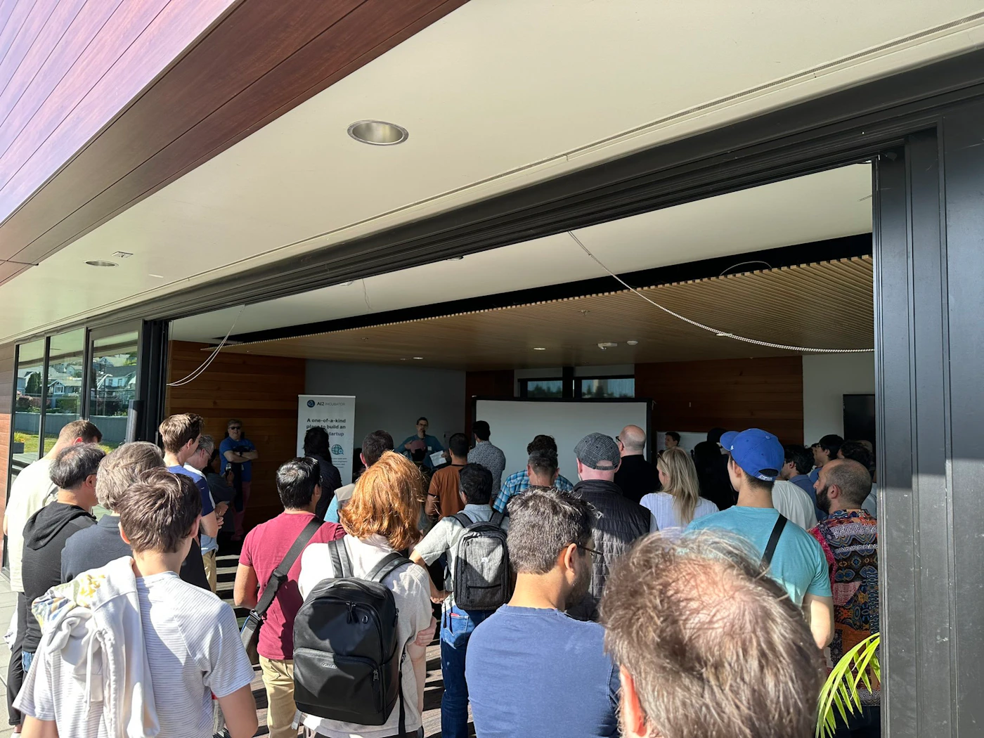 (Photo) A photo of a crowd of people gathered outside and partially inside a building, seemingly listening to a presentation. people | outdoor/indoor | crowd, presentation | candid Note: The image is a real-life captured scene of people in a specific setting, indicative of a photograph.