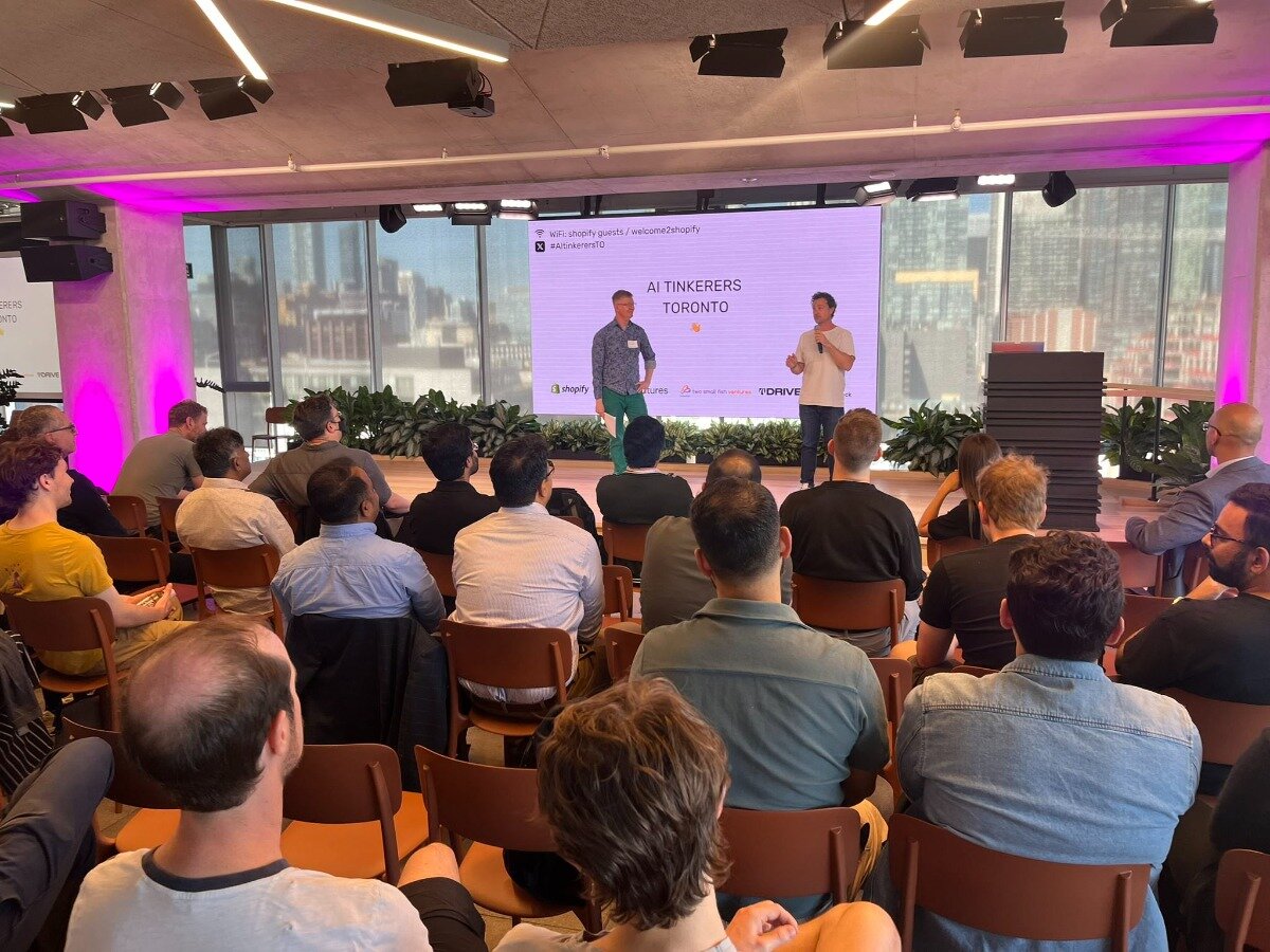 (Photo) A photograph captures an indoor event with an audience seated in rows, facing a stage where two presenters stand in front of a large screen. The screen displays event information and logos, and a city skyline is visible through large windows in the background. Text: wifi: shopify guests / welcome2shopify #AITinkerersTO AI TINKERERS TORONTO shopify TD DRIVE An event or conference with presenters and an audience. | Indoor, modern event space with large windows overlooking a city. | Multiple people (audience and presenters), a large projection screen, stage lighting, and a prominent city skyline in the background. | Candid, event photography. | Colors: #8B4513, #A9A9A9, #FF00FF, #FFFFFF, #4682B4 Note: This image is a direct capture of a real-world scene, showing people, an event setup, and a city view, which fits the definition of a photograph.