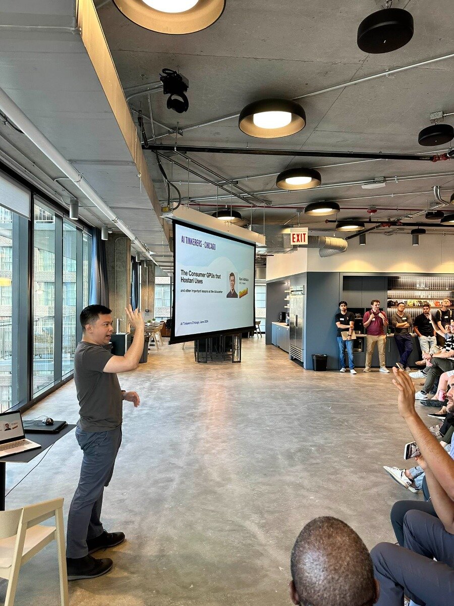(Photo) An indoor event showing a speaker presenting to an audience in a modern, industrial-style room with a projection screen displaying presentation slides. Text: AI TINKERERS - CHICAGO The Consumer GPUs that Hostari Uses and other important lessons at the datacenter Junie Abasolo Hostari AI Tinkers Chicago, June 22nd EXIT An indoor presentation or tech talk with a speaker and an audience. | Indoor, a large modern room with concrete floors, high ceilings with exposed pipes and lighting fixtures, and large windows. | A man presenting with a projection screen behind him, an audience seated and standing, a laptop on a table, and an 'EXIT' sign. | Candid, event photography. | Colors: #808080, #FFFFFF, #000000, #ADD8E6, #FF0000 Note: The image captures a real-world scene with people, a setting, and objects, characteristic of a photograph rather than an illustration, logo, or banner.