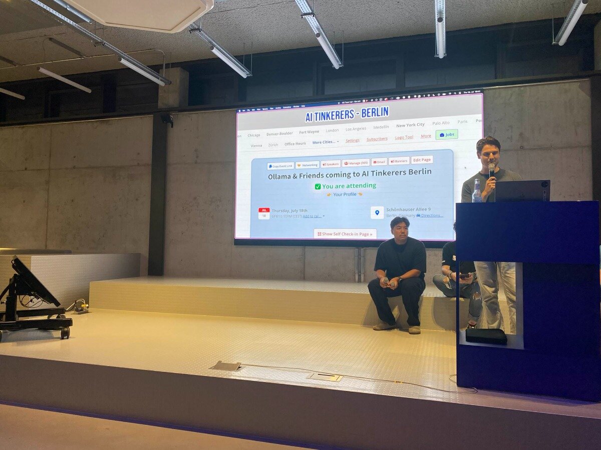 (Photo) A photograph captures an indoor tech event or presentation, featuring a speaker at a dark blue podium addressing an unseen audience, with a large screen behind him displaying event details for 'AI Tinkerers Berlin'. Another individual is seated on the white raised platform to the left of the speaker. Text: AI TINKERERS - BERLIN Chicago Denver-Boulder Fort Wayne London Los Angeles Medellin New York City Palo Alto Paris Zurich Office Hours More Cities... Join us! Subscribers Log In Tools More Jobs Reporting Speakers Manage data Email Banners Edit Page Ollama & Friends coming to AI Tinkerers Berlin ✓ You are attending Your Profile Thursday, July 18th Schönhauser Allee 9 Berlin, Germany Directions Show Self Check-in Page People, event, presentation, technology | Indoor, event space | Speaker at a podium, large projection screen, audience member on stage, modern architectural elements, concrete walls | Candid, event photography | Colors: #808080, #000080, #FFFFFF, #ADD8E6, #F5F5DC Note: The image is a direct capture of a real-world scene, showing people, an environment, and objects, which are characteristics of a photograph.