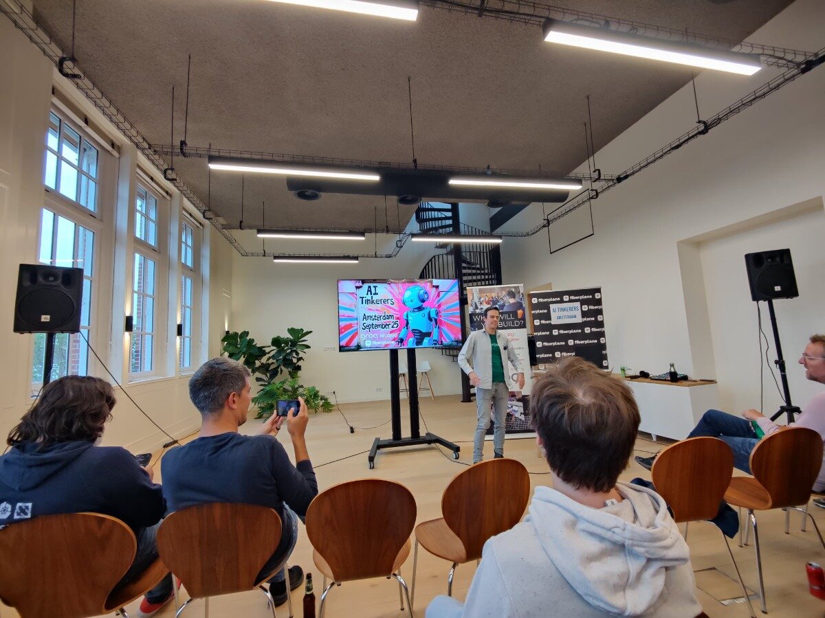 (Photo) A group of people are attending an indoor event, likely a tech meetup or presentation, with a speaker standing in front of a screen displaying a colorful slide. Several promotional banners are visible in the background. Text: AI Tinkerers Amsterdam September 25 WHY WILL BUILD? AI TINKERERS AMSTERDAM @Riverplane people attending an event | indoor, event space/conference room | a speaker presenting, an audience, a large display screen, promotional banners, and a spiral staircase in the background | candid, event photography | Colors: #EAEAEA, #A0522D, #000000, #FFFFFF, #FF007F, #008000 Note: The image is a photographic capture of a real-world event, depicting people, an indoor setting, and objects, which aligns with the definition of a 'photo'. It is not a graphic design element like a logo or banner, nor is it an illustration or handwriting.