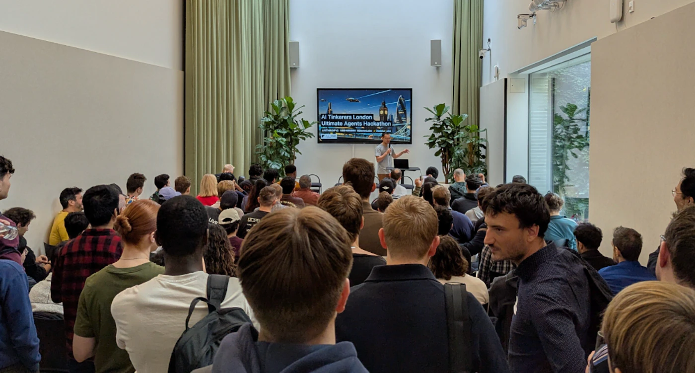(Photo) An indoor event shows a speaker presenting to a large audience, with many attendees visible from behind. A large screen displays text and an image of a futuristic London skyline, suggesting a tech-related event. Text: AI Tinkerers London Ultimate Agents Hackathon Date: [unreadable] / Time: [unreadable] / Location: [unreadable] / Register: [unreadable] A speaker giving a presentation to an audience at an event. | Indoor, likely an event hall or conference room. | A large group of people, a man speaking at the front, a large screen displaying event information and a futuristic city image, potted plants, and light green curtains. | Candid, event photography, wide shot. | Colors: #EAEAEA, #829C74, #2F2F2F, #D0D0D0, #000000 Note: The image is a direct capture of a real-world scene, featuring people, an indoor environment, and a presentation, which are characteristics of a photograph.