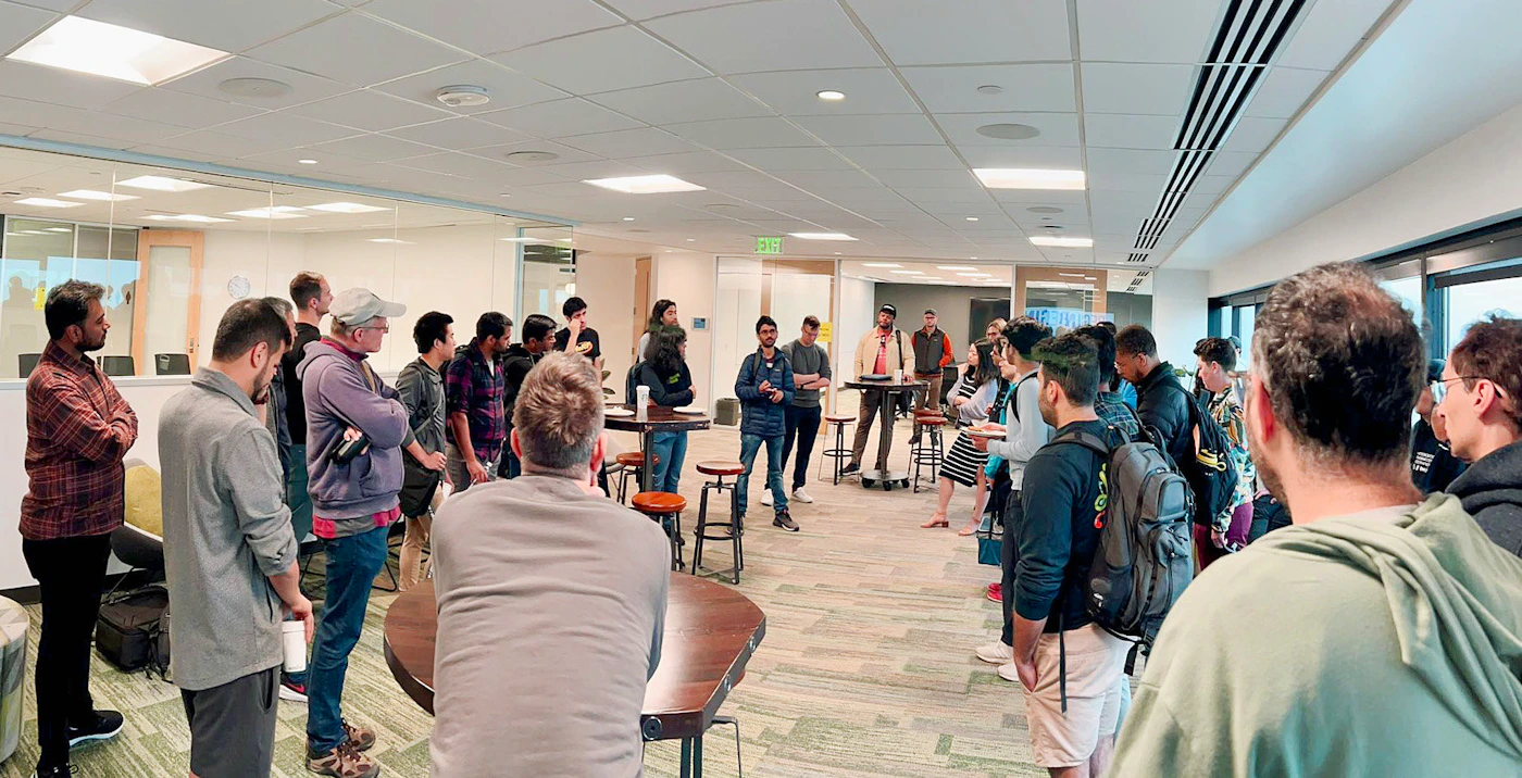 (Photo) A panoramic photo of a group of people gathered in an office space, some standing in a circle and others observing from the side. people | indoor | group of people, office setting | candid Note: The image is a real-life captured scene of people in an office environment, indicating a photograph. It's not a logo, illustration, banner, or handwriting.