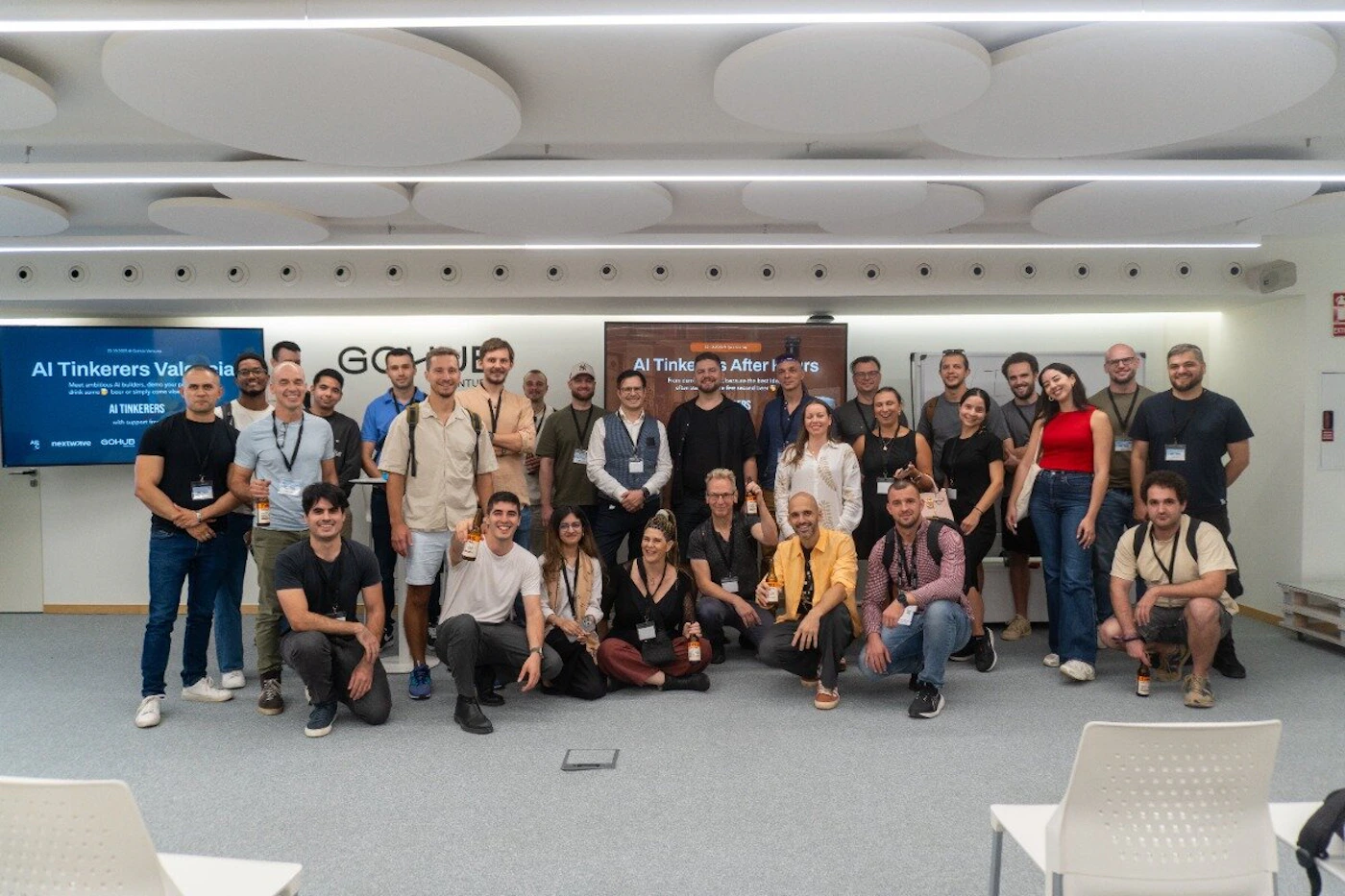 (Photo) A group of approximately 30 people posing for a group photo in a modern office or event space, likely after an AI-related meetup. Text: AI Tinkerers Valencia Meet ambitious AI builders, demo your projects, or simply come chat AI TINKERERS with support from NEXTSCENE GOHUB AI Tinkerers After Hours Enjoy some drinks... Check out the food and... after your [unreadable] in the second floor group of people | indoor office or event space | multiple people, digital screens with event information, modern acoustic ceiling panels | group portrait | Colors: #E0E0E0, #1A1A1A, #004080, #C0392B Note: This is a real-life photograph capturing a group of individuals in a physical setting.